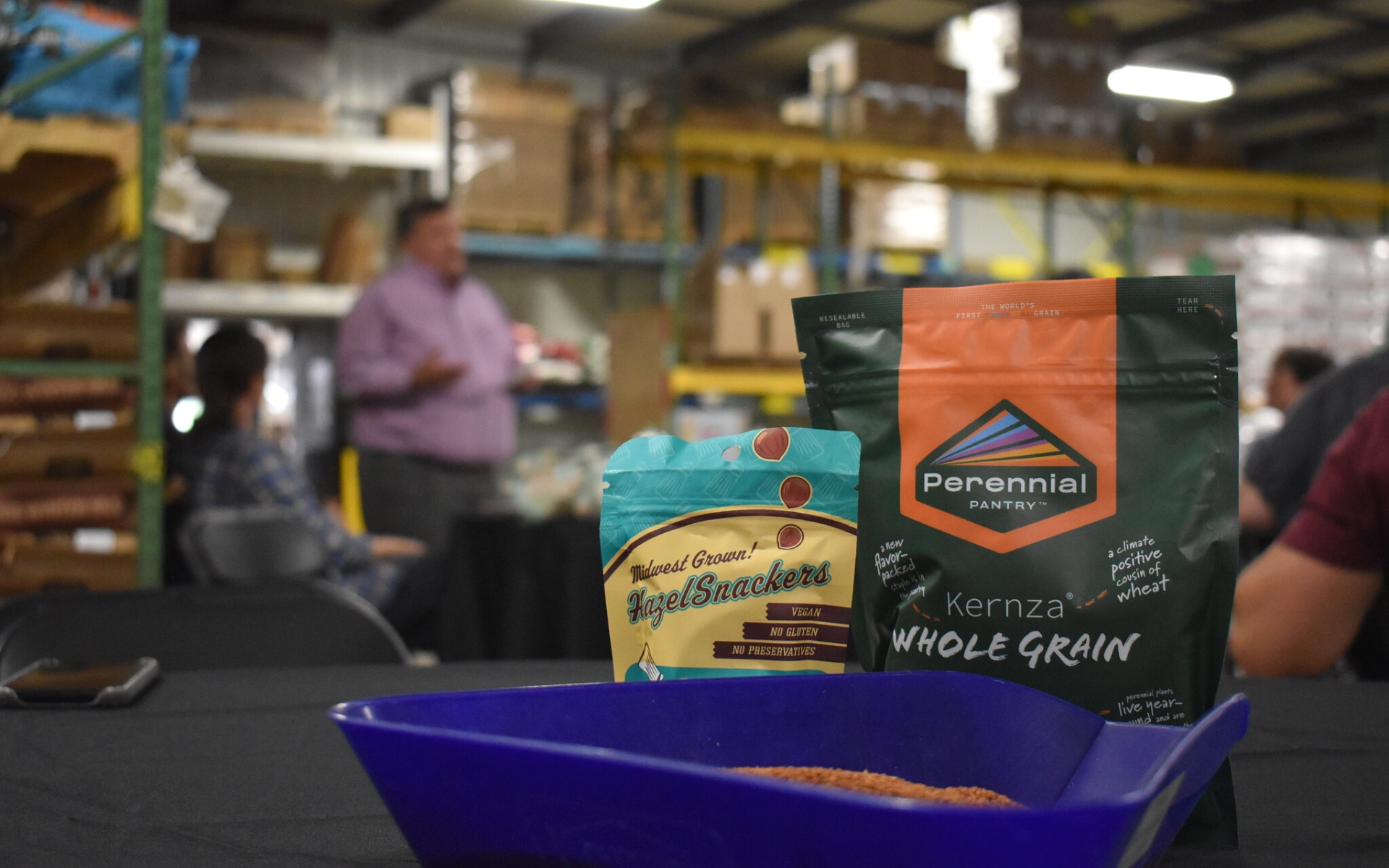 Two packets of food snacks made with clean-water crops sit on a table in the foreground. In the blurred background, you can make out warehouse shelves and individuals talking.