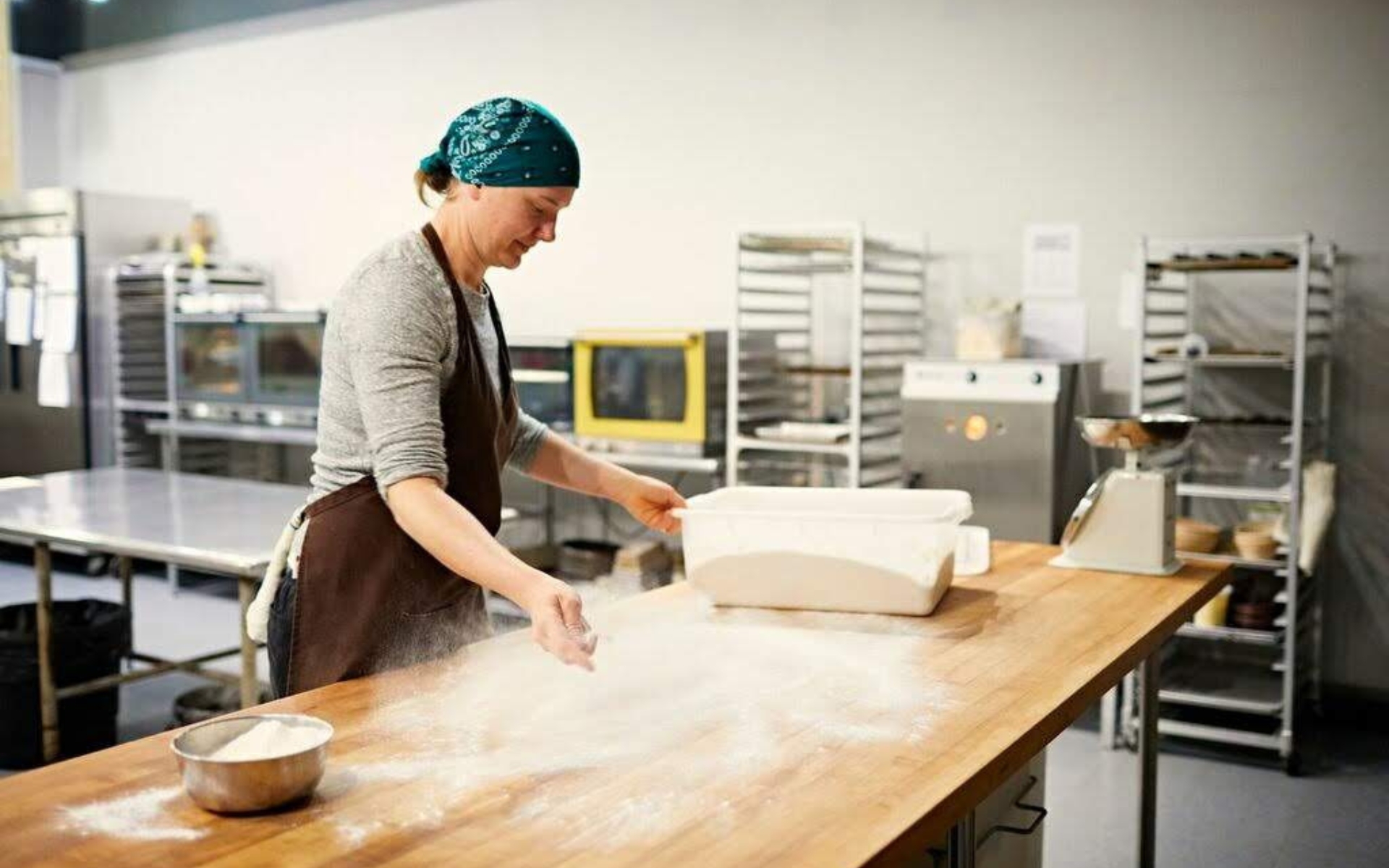 A baker at a large table, spreading flour as dough sits waiting to the side. 