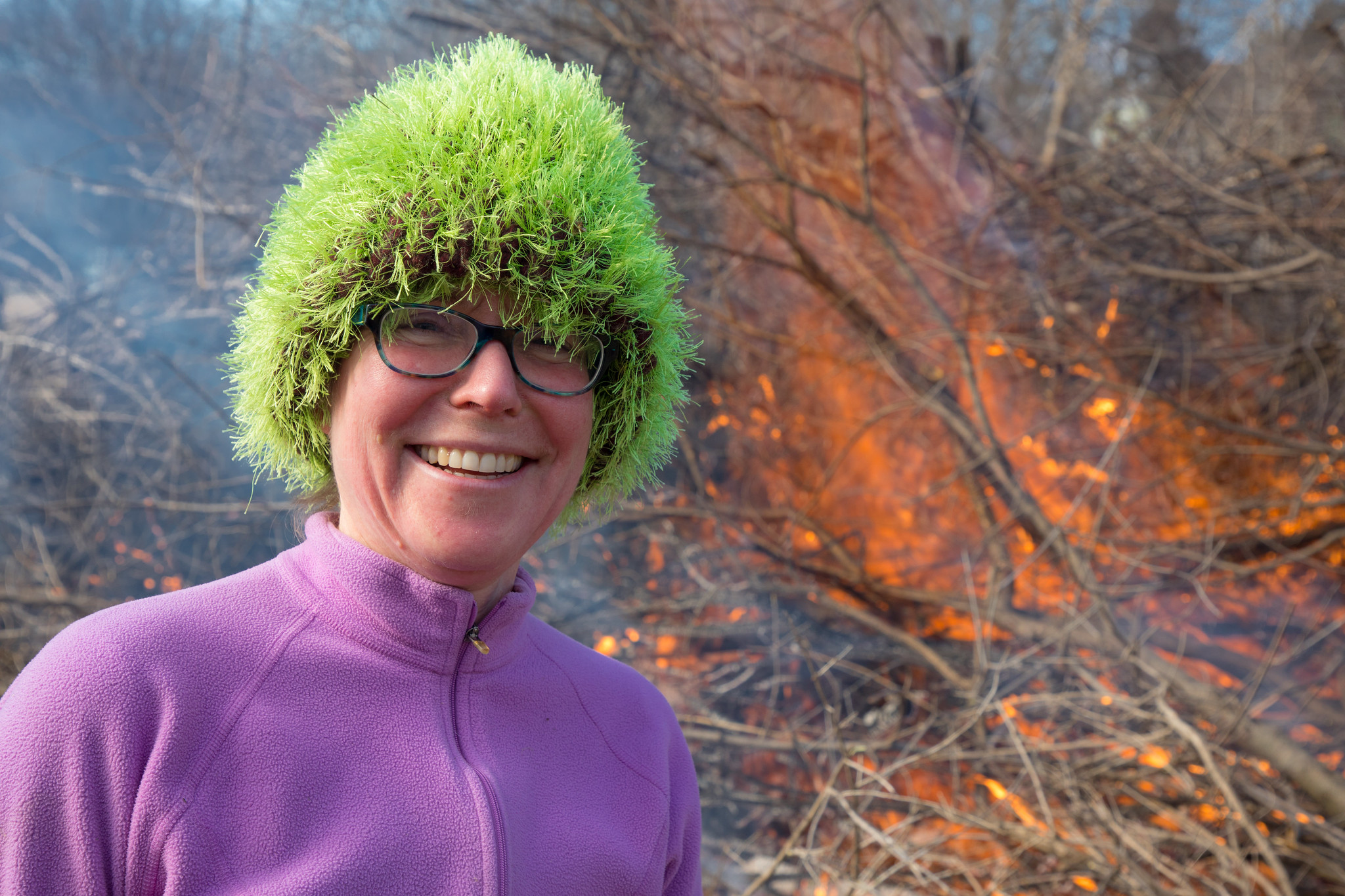 A volunteer in a fuzzy green hat smiles in front of a buckthorn bonfire