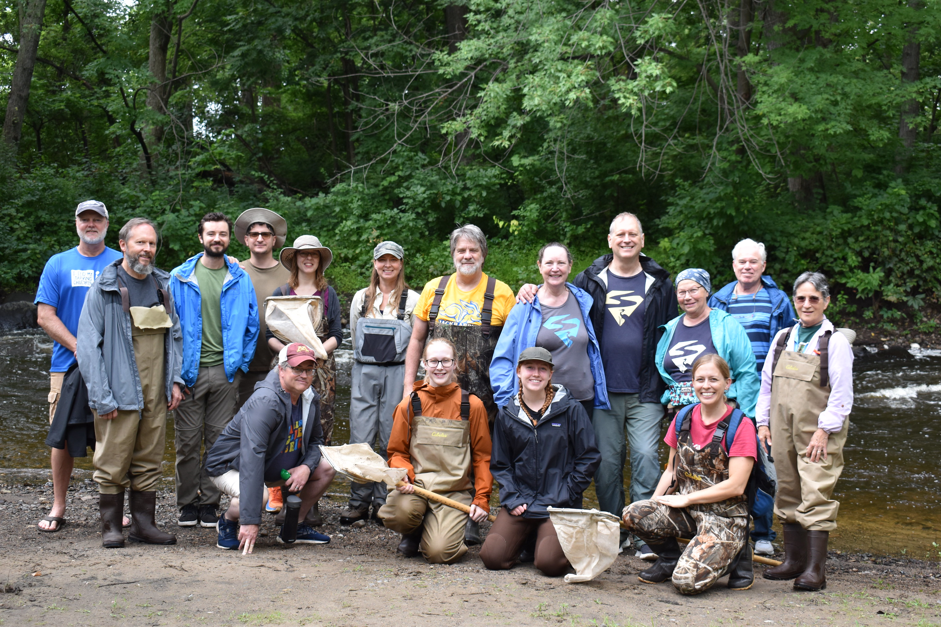 16 volunteers from the Stream Health Evaluation Program pose in front of a stream. Many of them are wearing waders and are smiling.