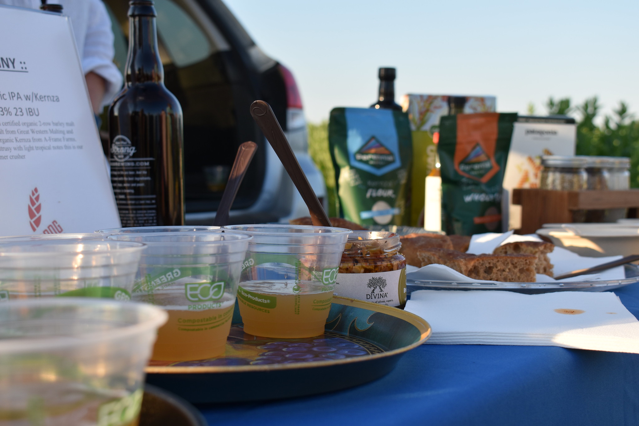 Products made using clean-water crops displayed on a table. Biodegradable cups are half-full with beer. A spoon sticks out of a small jar. Flour in its package is in the background.