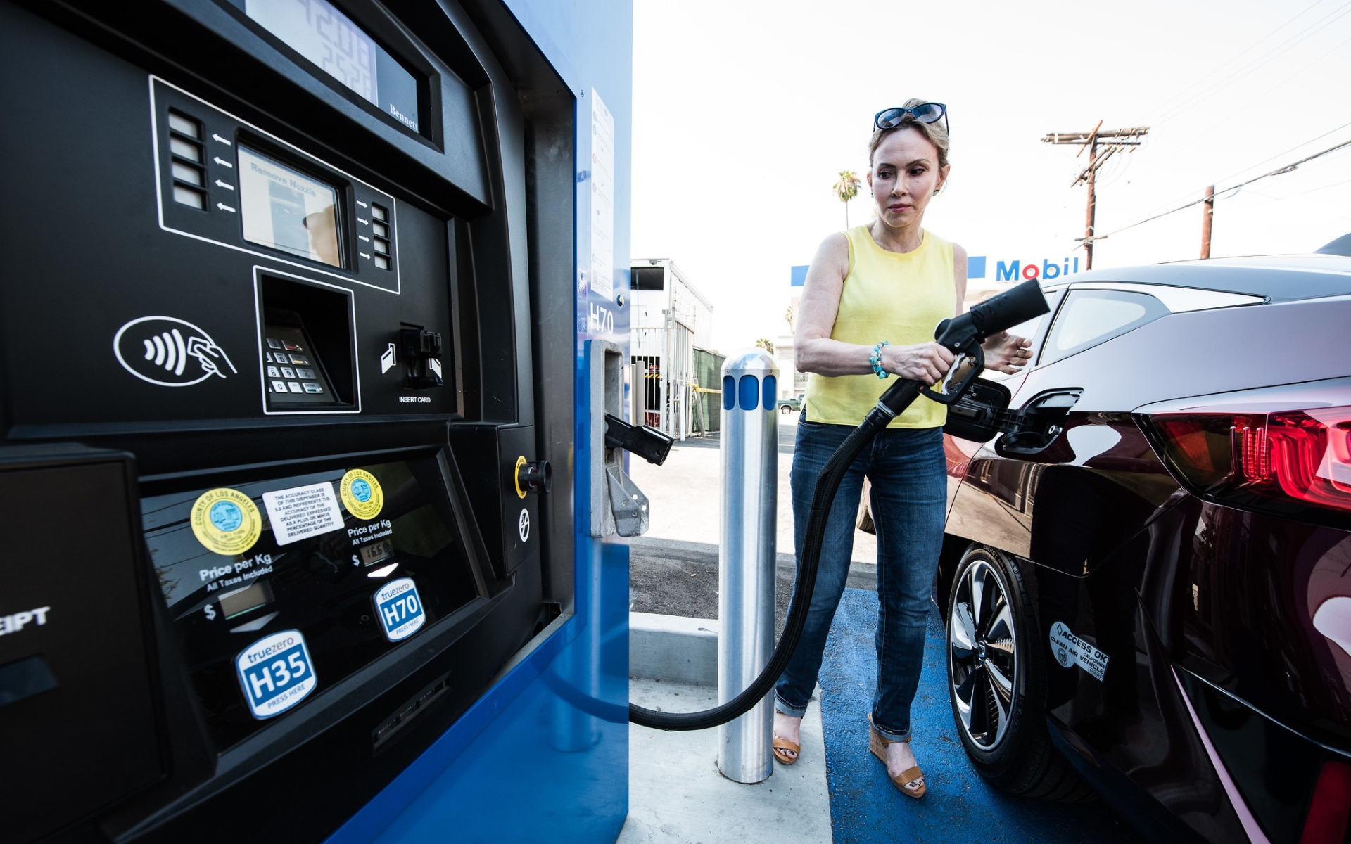 A driver at a hydrogen fueling station, filling up her fuel cell vehicle. She is holding a pump that appears similar to a traditional gas station.