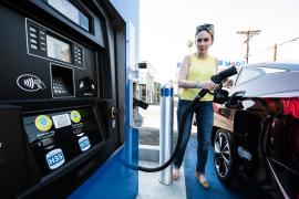 A driver at a hydrogen fueling station, filling up her fuel cell vehicle. She is holding a pump that appears similar to a traditional gas station.