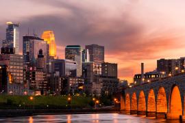 The Minneapolis skyline, reflective against the orange evening sky with the Mississippi River in the foreground.
