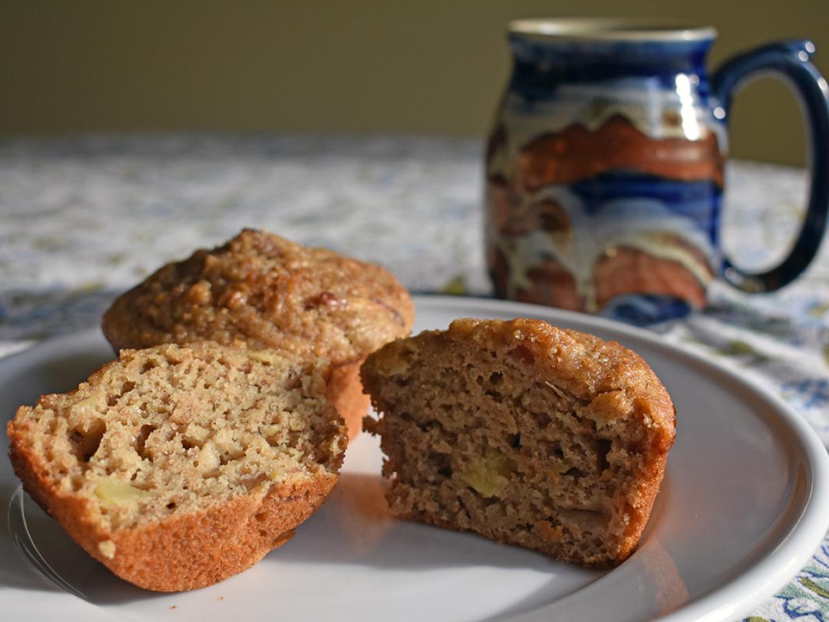 A recently baked cinnamon apple Kernza muffin is split apart on a plate. A mug can be seen on the table just behind it.